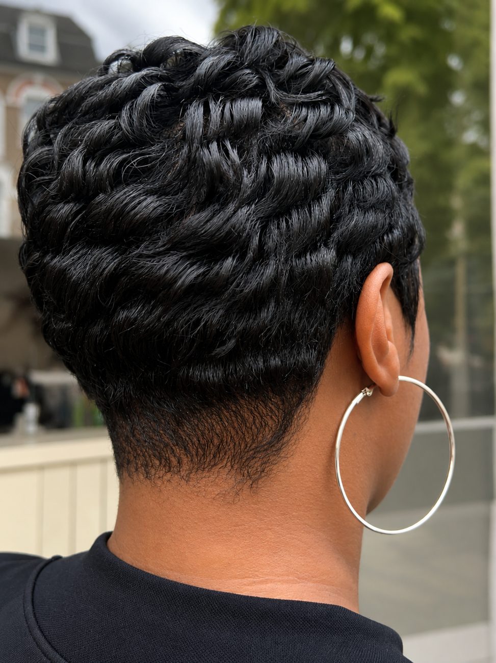 Back view of a sculpted short-hair restyle with defined curls and a silver hoop earring, showing the nape and finish detail